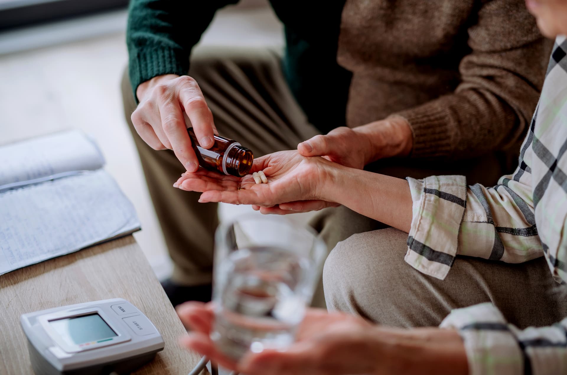 Two people seated closely; one pours pills from a brown bottle into the other's hand, who also holds a glass of water. 