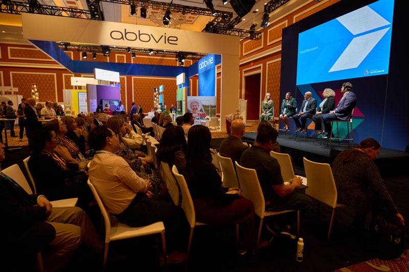 Audience seated at a conference watches a panel discussion on a stage near an AbbVie booth, with large screens and expo displays in the background.