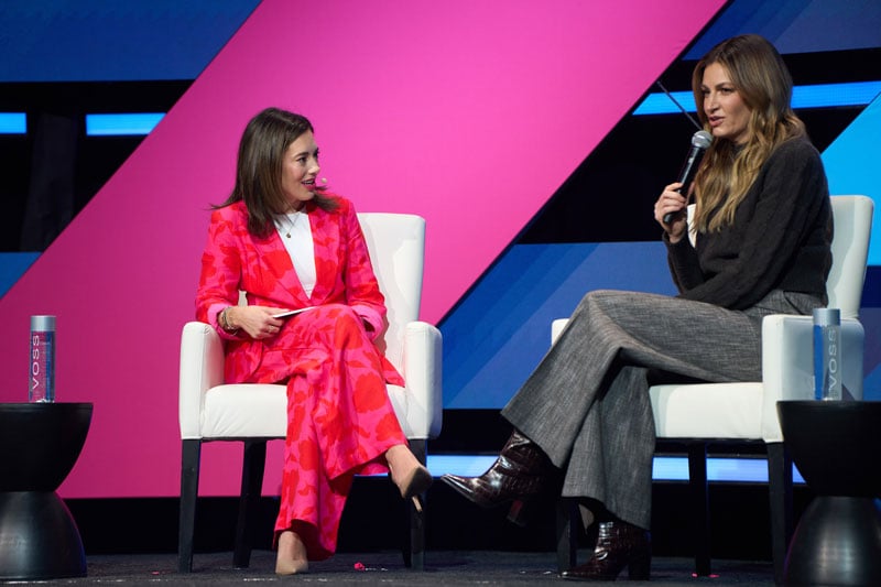 Two women sit in white chairs on a stage, one holding a microphone, engaged in a conversation against a bold pink and blue geometric backdrop.