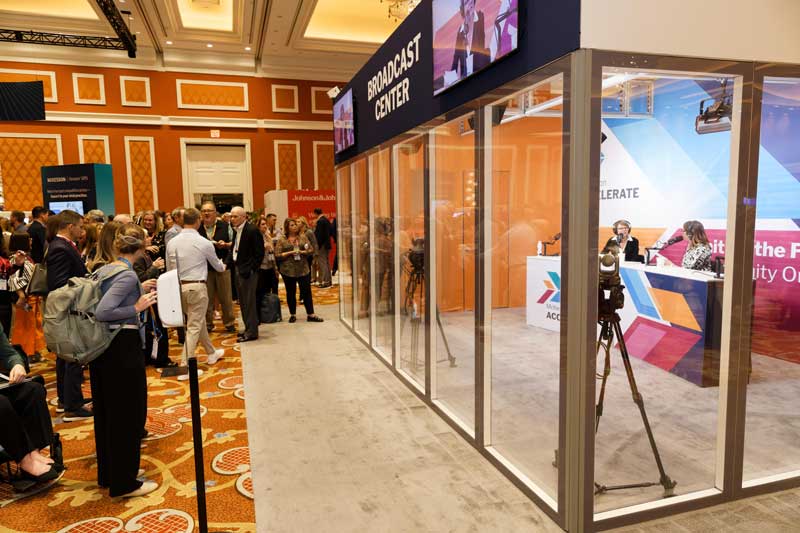 Crowd watches a live broadcast inside a glass studio at a conference hall, with cameras, hosts at a desk, and signage overhead.