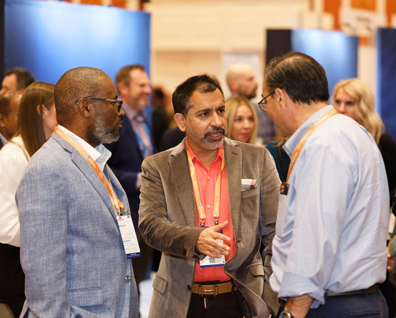 Three conference attendees wearing badges talk in a busy expo hall, gesturing as they network among crowds and display booths.