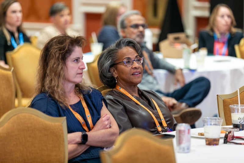 Conference attendees sit at round tables listening to a session, with two women in the foreground wearing badges and taking notes.