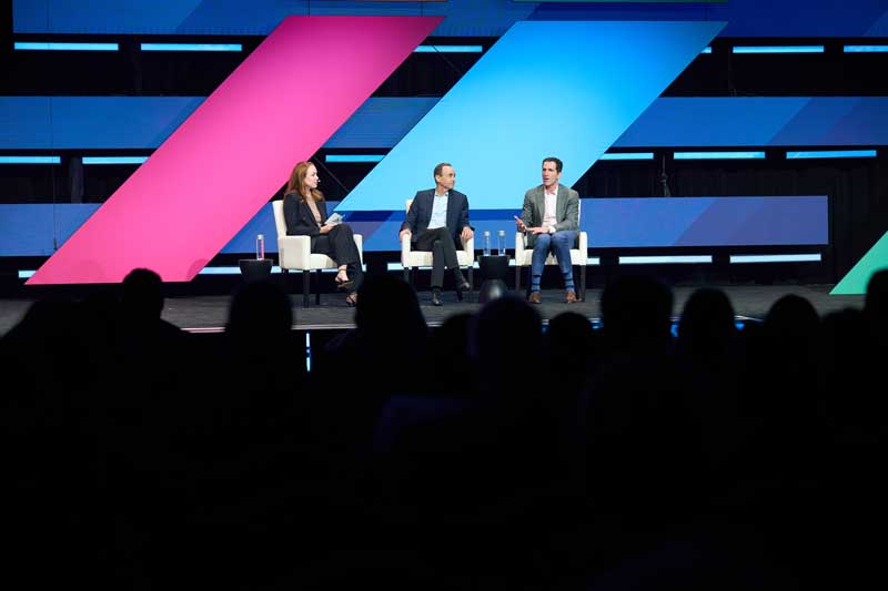Three speakers sit in white chairs on a stage, speaking to an audience, with large pink and blue geometric panels behind them.