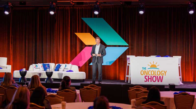 Speaker stands on a conference stage with colorful geometric backdrop, seated audience in front, and a set branded “The Oncology Show” to the side.