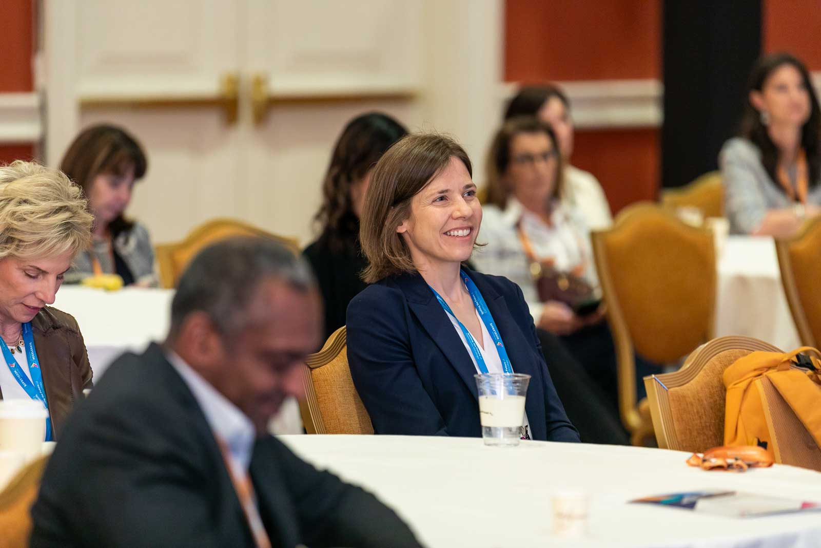 Attendees seated at round tables during a conference session, wearing badges, with notebooks and drinks on tables.