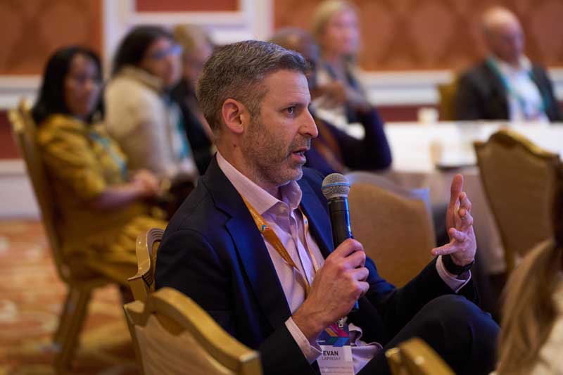 A man seated at a conference holds a microphone and gestures while speaking; attendees sit behind him in a hotel ballroom.