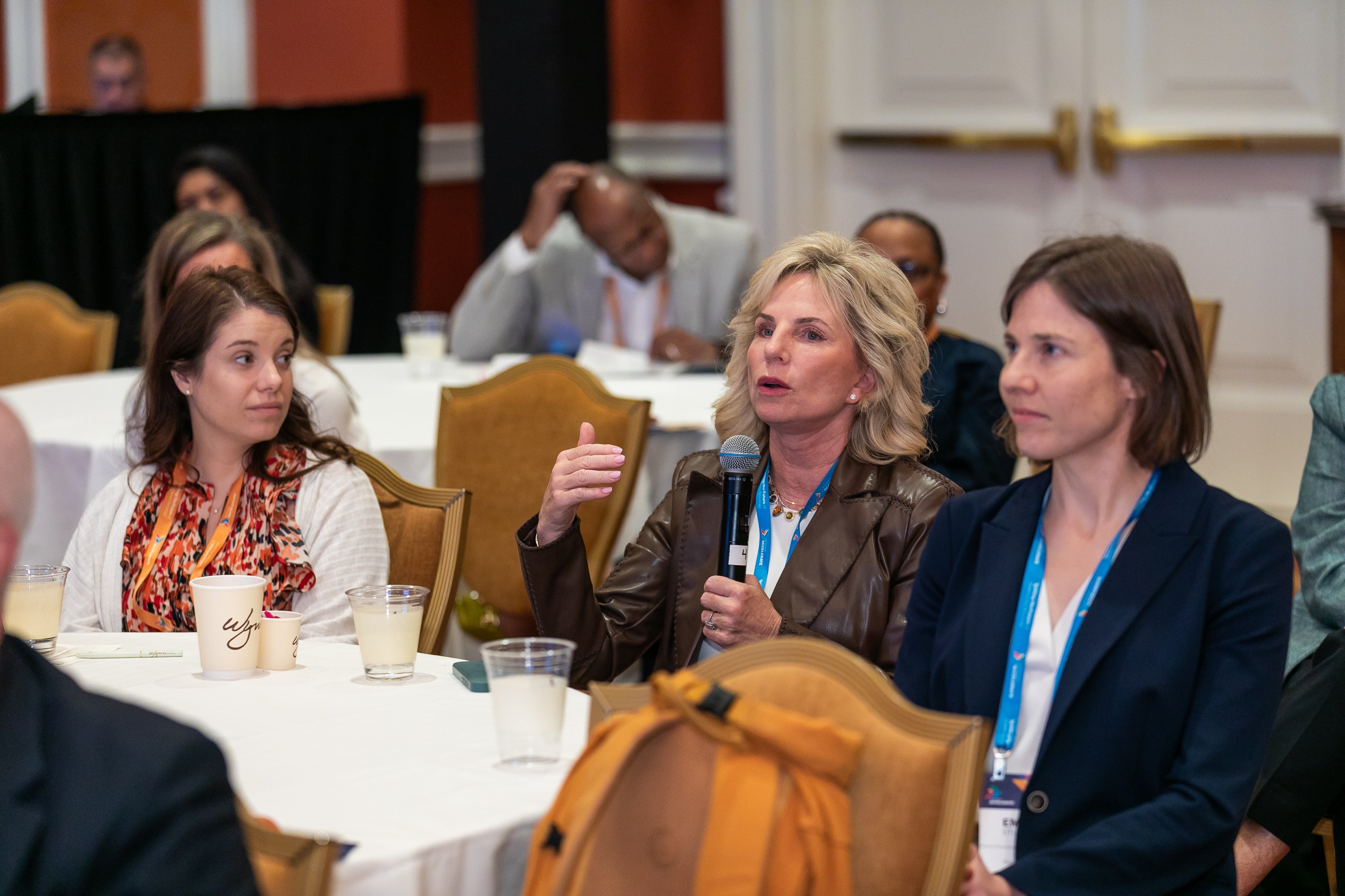 Panel discussion at a conference: a woman speaks into a microphone at a round table while attendees listen and take notes.