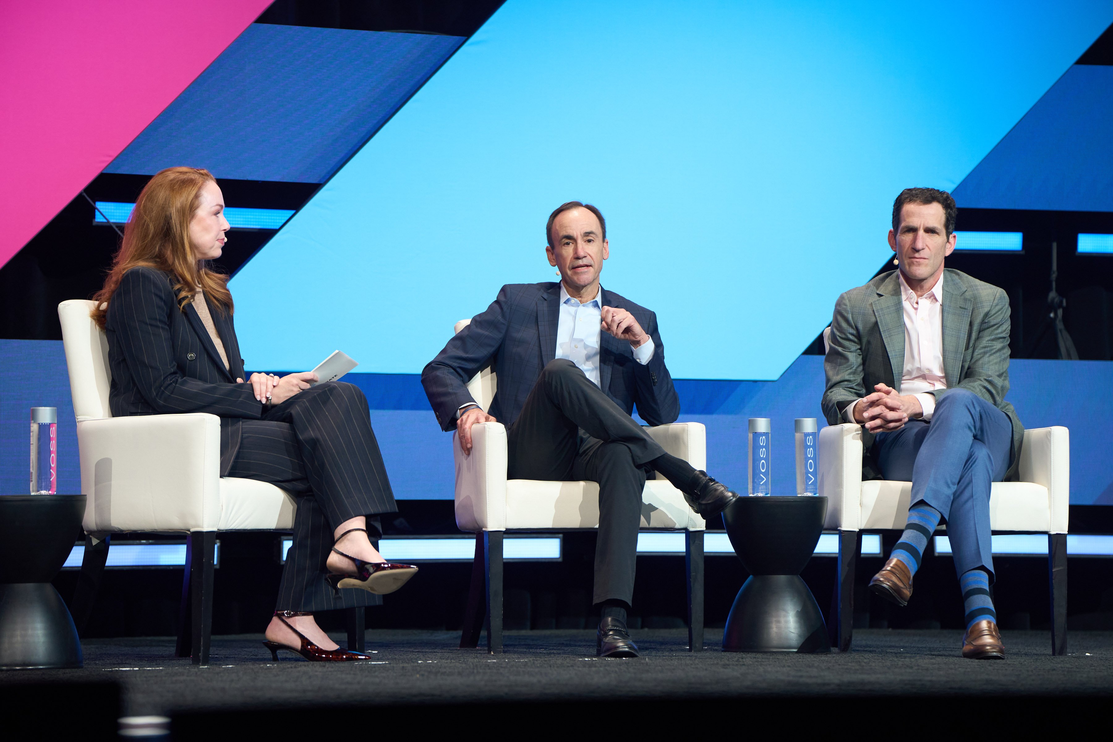 Three panelists seated on white chairs on a conference stage, engaged in discussion, with a large blue and pink geometric backdrop behind them.