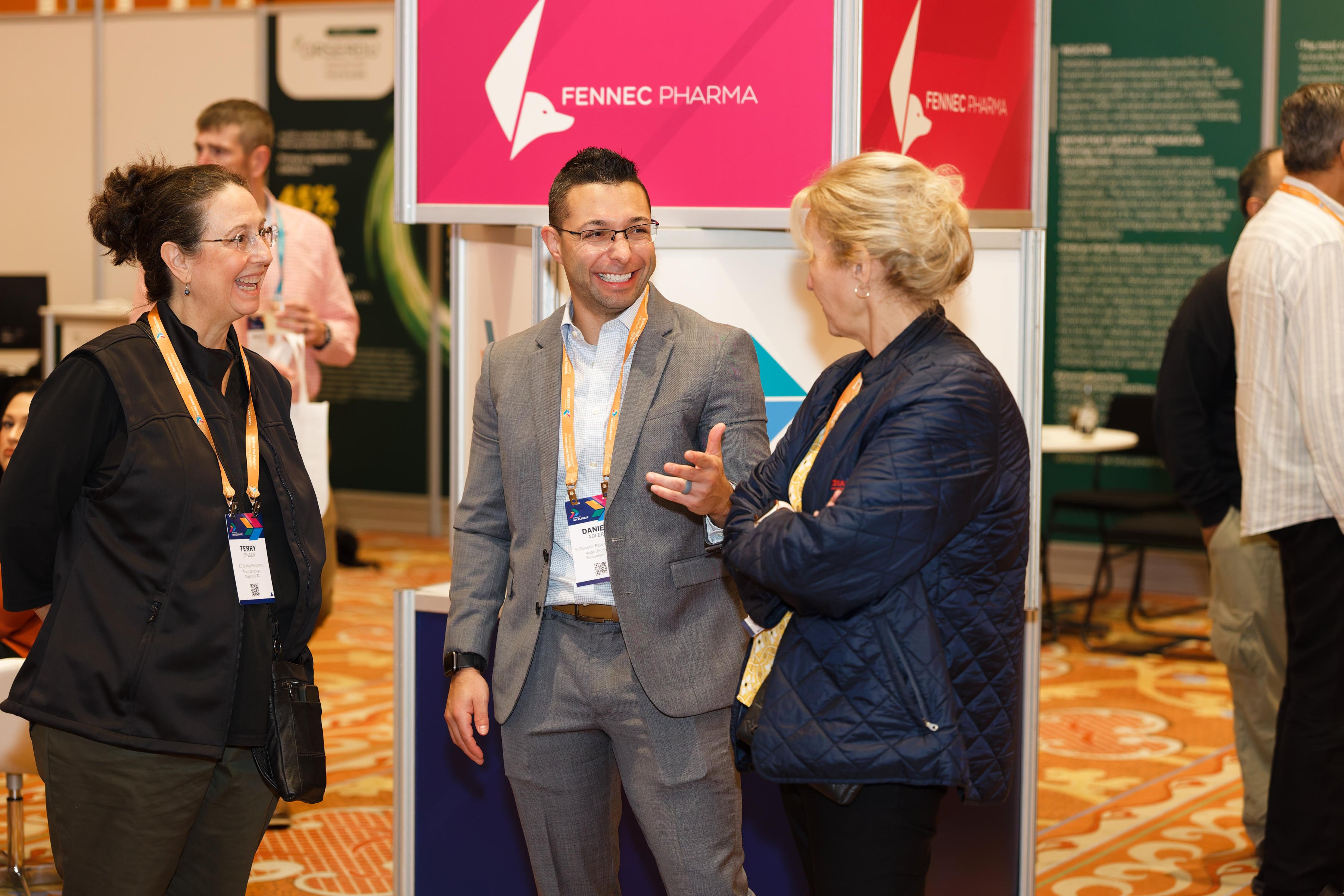 Three conference attendees wearing badges converse at a Fennec Pharma booth on a trade show floor.
