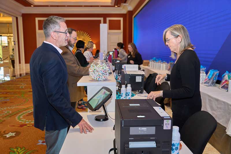 Two people talk across a registration counter at a conference, with badges, water bottles, and check-in equipment in a hotel ballroom.