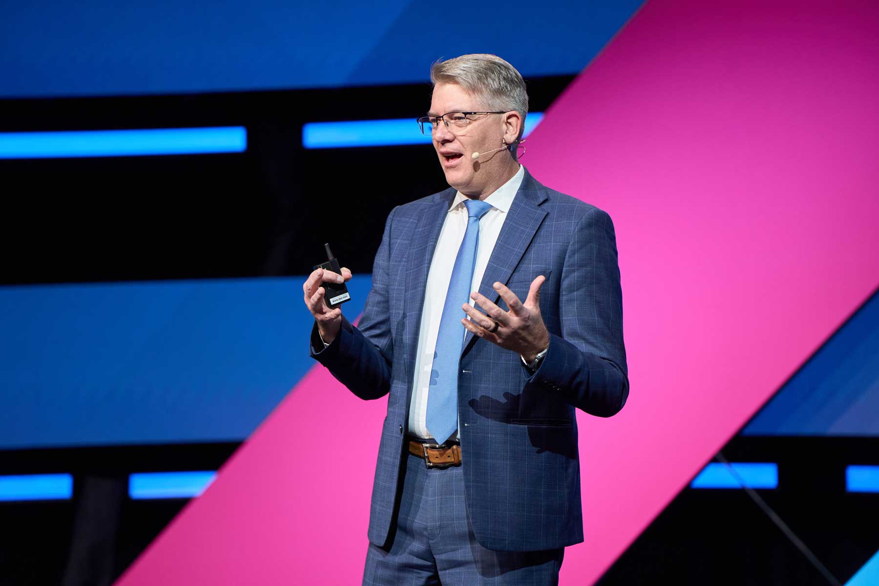 Person in a blue suit and tie speaking on stage, holding a remote, with a large pink and blue geometric backdrop behind them.
